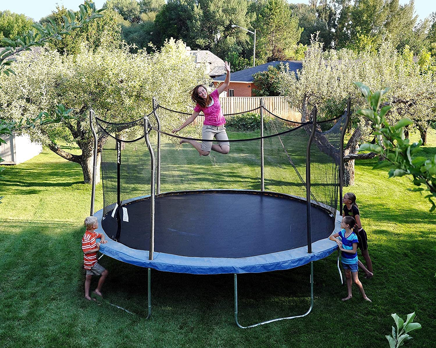 Backyard trampoline fun: a girl jumping while other children watch, surrounded by trees and green lawn